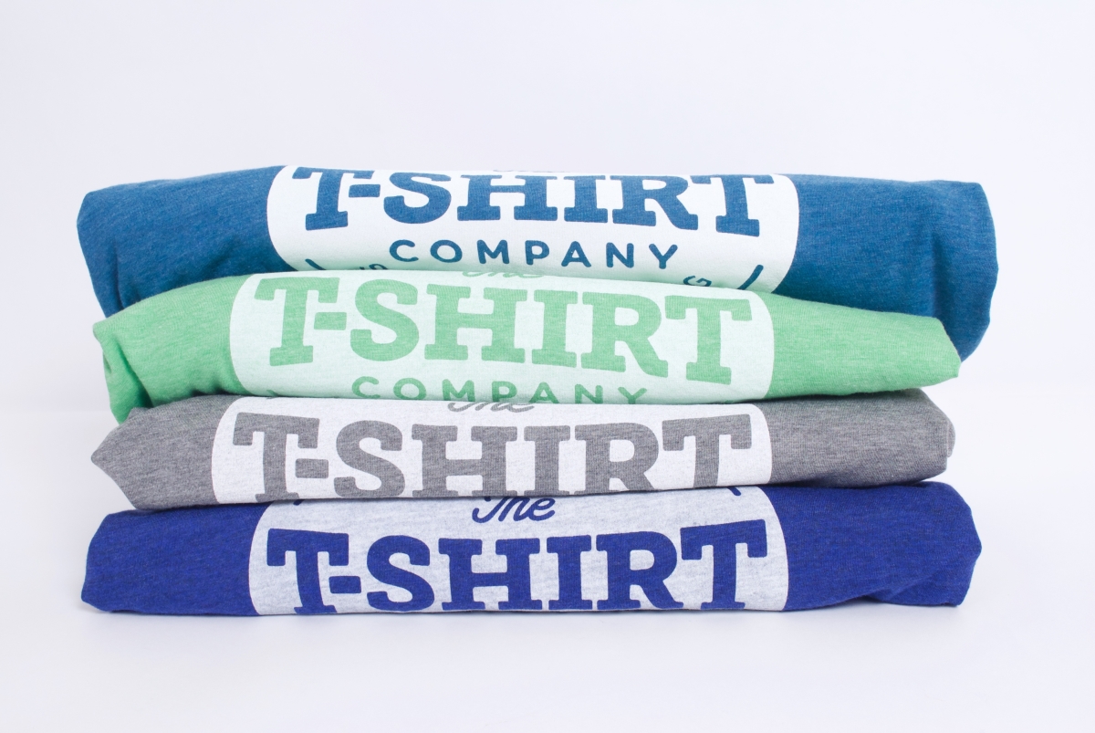 4 triblend tees in green, grey,  aqua and true royal blue stacked on top of each other with the t-shirt company logo printed in white discharge waterbased ink visible on each Bella and Canvas t-shirt