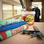 Three screen-printed gym t-shirts with a pot of the tshirt company red discharge ink and printer's tool on wooden surface, window and various tees in background